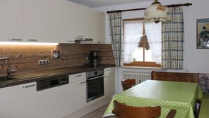 A compact kitchen with a dishwasher, oven, and a coffee maker. There is a dining table with chairs and a window with curtains. - Grünau on the ground floor (Schönau am Königssee)