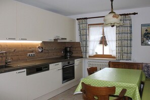 A compact kitchen with a dishwasher, oven, and a coffee maker. There is a dining table with chairs and a window with curtains. - Grünau on the ground floor (Schönau am Königssee)