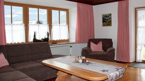 A living room with a sofa, armchair, and a coffee table. There are windows with curtains, a radiator, and a painting on the wall. - Grünau on the ground floor (Schönau am Königssee)
