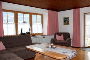 A living room with a sofa, armchair, and a coffee table. There are windows with curtains, a radiator, and a painting on the wall. - Grünau on the ground floor (Schönau am Königssee)