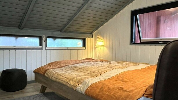 Uma cama de solteiro com cabeceira, uma janela com persianas, uma luminária fixada na parede e um banquinho redondo.