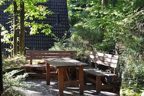 Un banc et une table en bois sont installés dans un jardin où la végétation est luxuriante, avec un mur de pierre en arrière-plan.