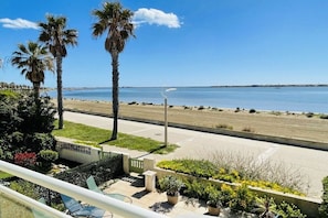 A balcony with a view of the beach, palm trees, and a clear sky. - La Baie du Levant 12 (Gruissan)