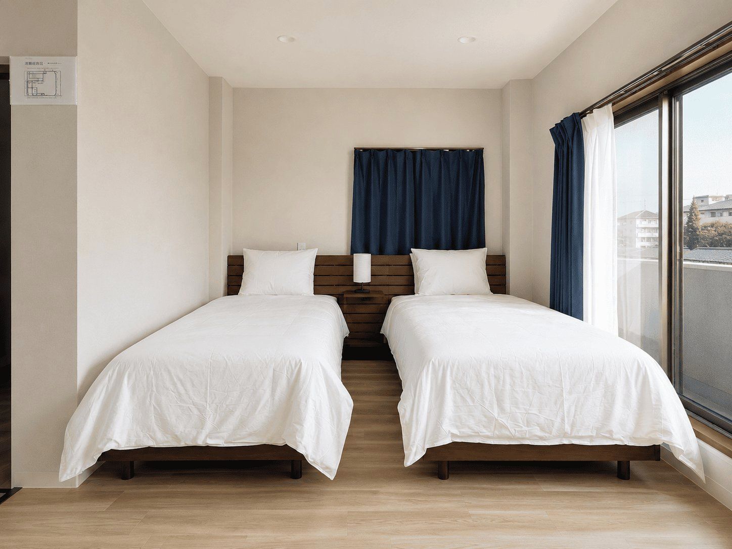 A double room with two beds, a wooden headboard, a large window with curtains, and a view of buildings outside.
