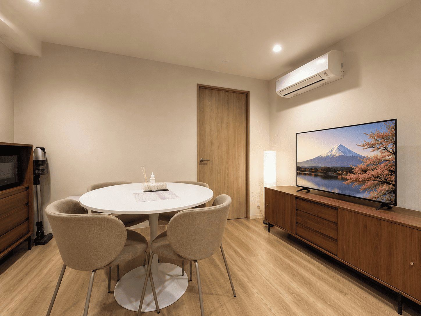 A hotel room with a round dining table, four chairs, a flat-screen TV mounted on a wooden cabinet, and an air conditioning unit on the wall.
