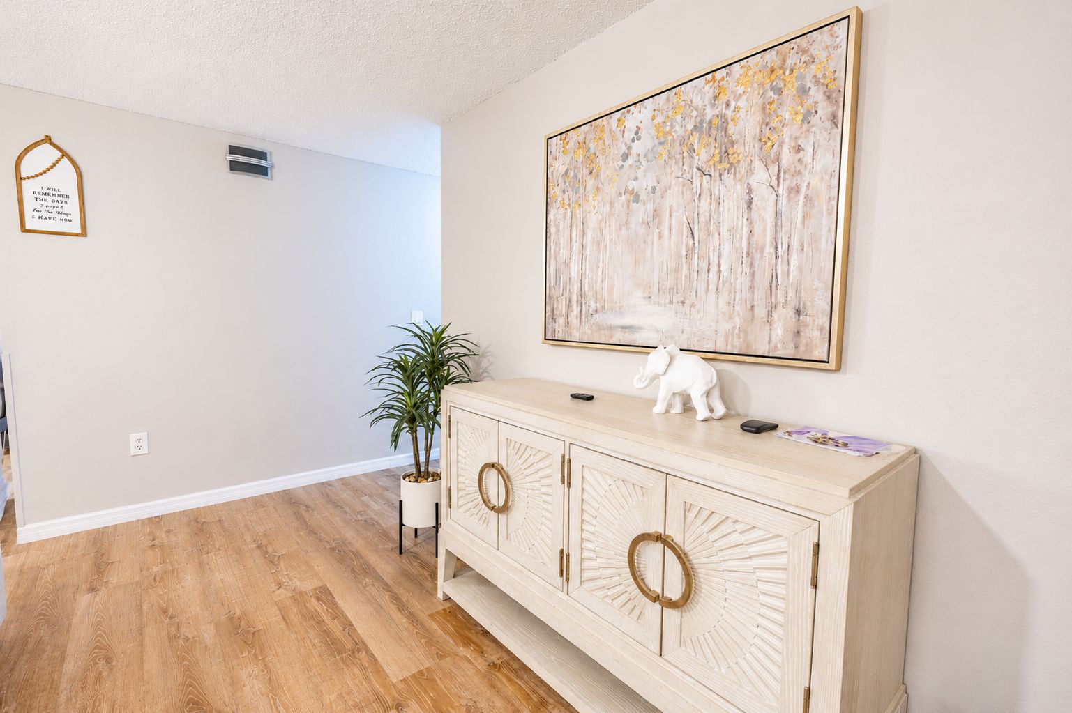 Een hal met een houten vloer, een witte muur, een kast met decoratieve knoppen, een kamerplant en een ingelijst inspirerend citaat.