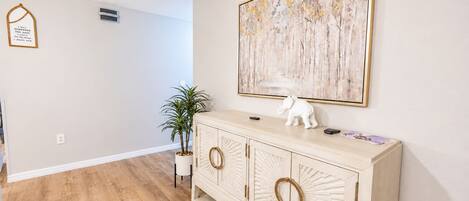 Een hal met een houten vloer, een witte muur, een kast met decoratieve knoppen, een kamerplant en een ingelijst inspirerend citaat.