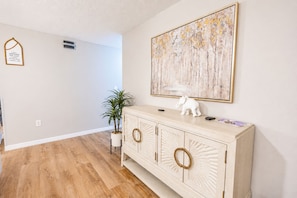 A hallway with a wooden floor, a white wall, a cabinet with decorative handles, a potted plant, and a framed inspirational quote. - Christmas Magic Starts Here/Book now and unwrap a special stay surprise! (BRADENTON)