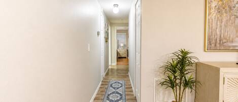 A hallway with a wooden floor, a patterned rug, a potted plant, and a piece of wall art.