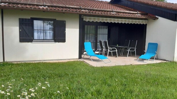 A house with a tiled roof, a covered patio with blue chairs and a table, and a lawn with white flowers. - Holiday home "Hochplatte" (Lechbruck)