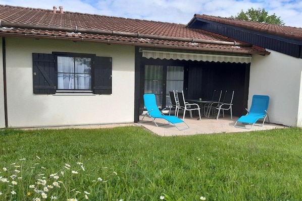 Uma casa com telhado de telhas, varanda coberta com cadeiras azuis e uma mesa, e um gramado com flores brancas.