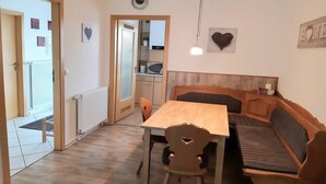A dining area with a wooden table and bench seating, a kitchen in the background, and a door leading to an outdoor area. - Holiday home "Hochplatte" (Lechbruck)