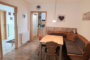 Área de refeições com mesa de madeira e bancos, cozinha ao fundo e porta que dá para área externa.