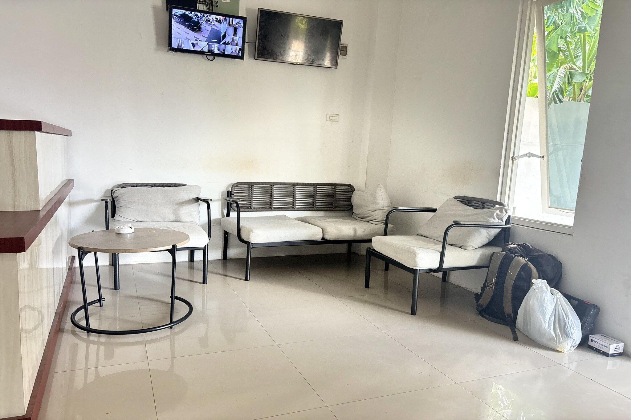 A room with a sofa, two chairs, a small table, a TV mounted on the wall, and a window with a view of greenery.