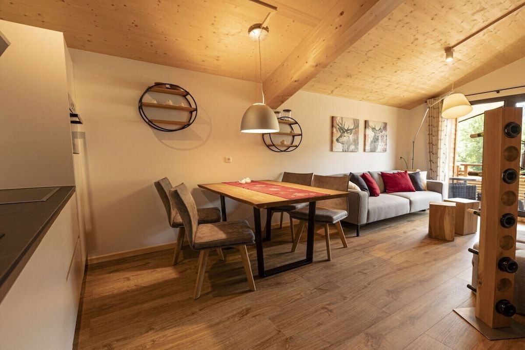 A spacious living area with a wooden ceiling, a dining table with chairs, a sofa with cushions, and a large window.