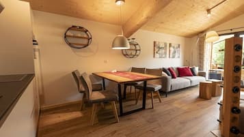 A spacious living area with a wooden ceiling, a dining table with chairs, a sofa with cushions, and a large window.