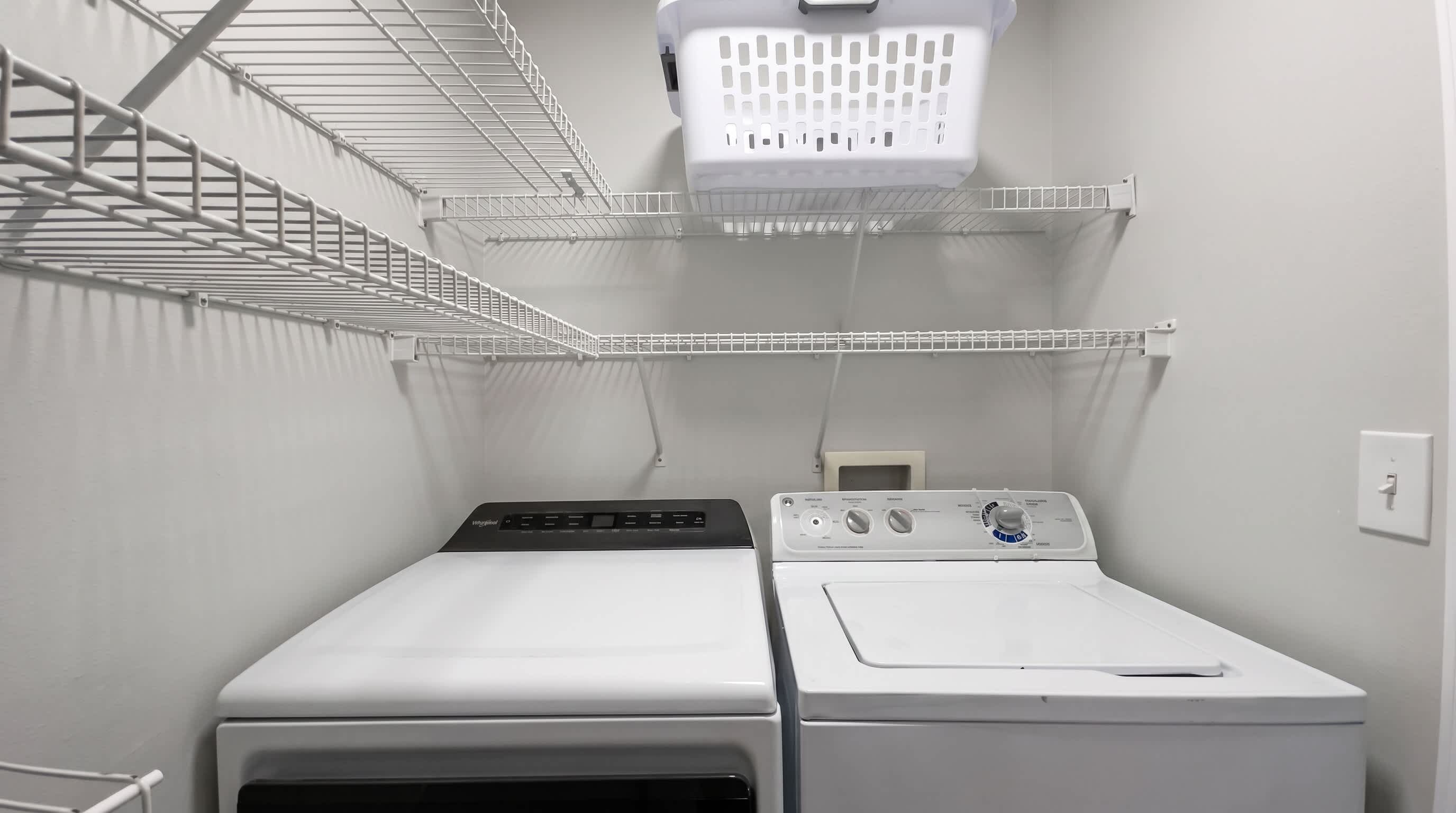 Une buanderie équipée de deux machines à laver et deux sèche-linge superposés, d’étagères en métal et d’un panier à linge suspendu.