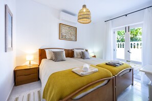 A double bed with a wooden headboard, a bedside table with a lamp, a hanging light fixture, and a window with a view of greenery.