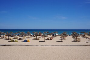 Une plage bordée de rangées de chaises longues et d’ombrelles en chaume, une promenade en bois et l’océan en toile de fond.