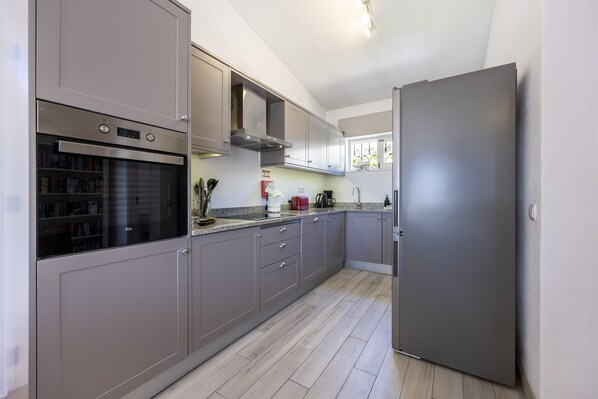 A modern kitchen with grey cabinets, a built-in oven, a stainless steel refrigerator, and a window providing natural light.