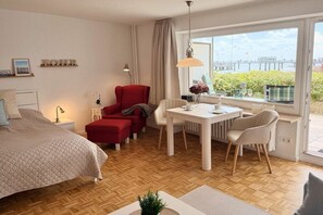 A modern hotel room with a bed, a red armchair, a dining table with chairs, and a view of a bridge through large windows. - Holiday apartment with 1 bedroom (Wyk auf Föhr)
