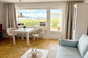 Ein Wohnzimmer mit einer Couch, einem Esstisch mit Stühlen, Blick ins Freie und einem fenster mit Vorhängen.