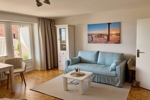 A living room with a light blue sofa, a small white coffee table, and a painting of a sunset on the wall. - Holiday apartment with 1 bedroom (Wyk auf Föhr)