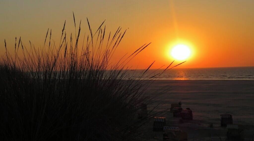 A sunset over a beach with grass in the foreground and a few chairs visible.