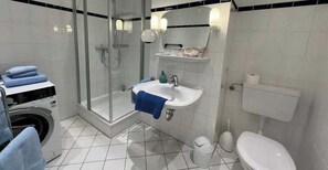 A bathroom with a glass-enclosed shower, a sink with a towel, a toilet, and a washing machine. - Apartment 260 - Dünenliebe (Juist)