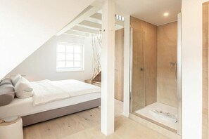A modern bedroom with a sloped ceiling, a bed with pillows, a bedside table, and a walk-in shower with a glass door. - 238 - Holiday home Watt (Juist)
