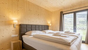 Un dormitorio con techo de madera, una cama con ropa blanca, dos lámparas de noche y vistas a un paisaje a través de una ventana.
