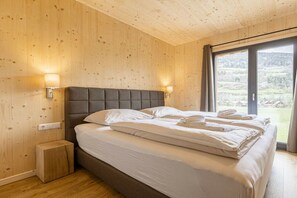 Une chambre avec un plafond en bois, un lit recouvert de draps blancs, deux lampes de chevet et une vue sur un paysage par la fenêtre.