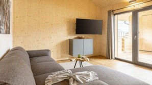 Una sala de estar moderna con un sofá gris, un televisor de pantalla plana montado en la pared, un mueble bajo y un gran ventanal corredero que da a un espacio exterior.