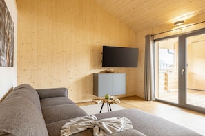 Un salon moderne avec un canapé gris, un téléviseur à écran plat fixé au mur, un petit meuble et une porte-fenêtre coulissante donnant sur un espace extérieur.
