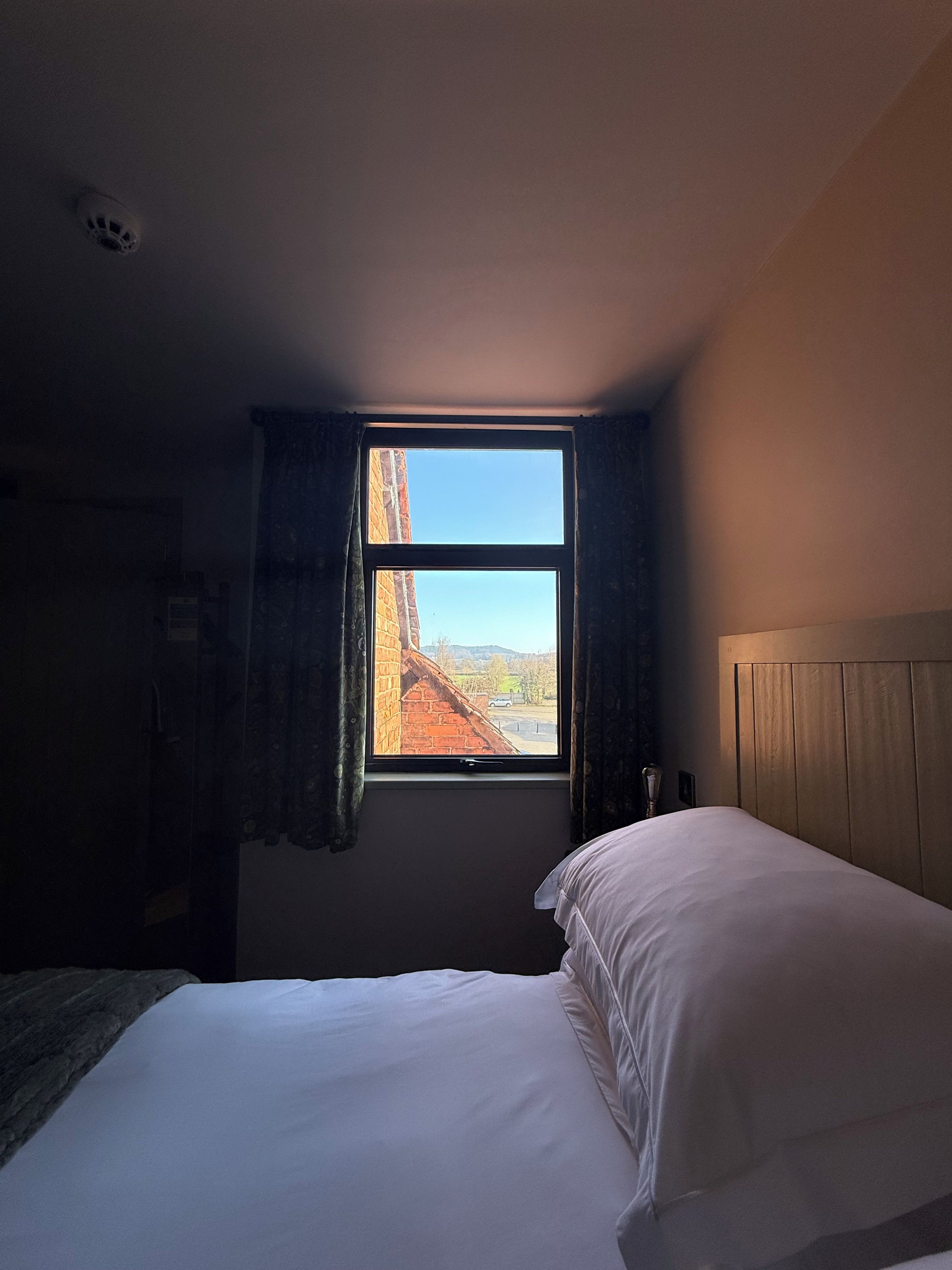 Une chambre avec un lit simple, une fenêtre avec des rideaux et une vue sur un bâtiment en briques à l’extérieur.