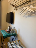 A room with a television mounted on the wall, a table with various items, a chair, and a coat rack with hangers.