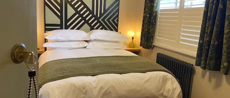 A bedroom with a bed, a bedside table with a lamp, a radiator, and a window with curtains.