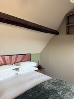 A bedroom with a sloped ceiling, a bed with white bedding, a wooden headboard, and a small bedside table with a lamp.