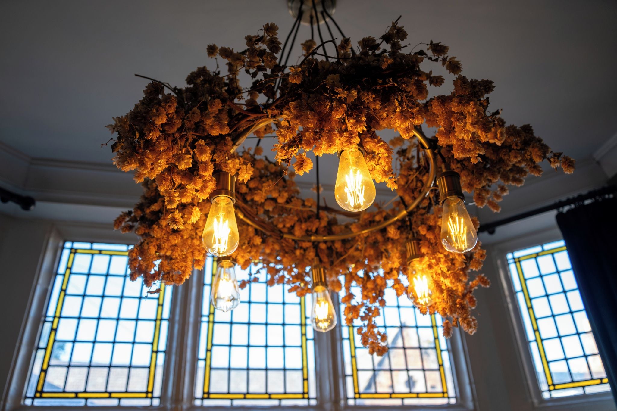 Un candelabro con múltiples bombillas, rodeado de flores secas, en una habitación con ventanas amplias.