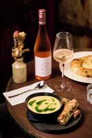 Un tavolo da pranzo con una ciotola di zuppa, una bottiglia di vino, un bicchiere di vino, un piatto con del pane e un vaso con fiori secchi.