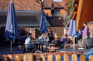 An outdoor dining area with tables and chairs, umbrellas, and a brick building in the background.