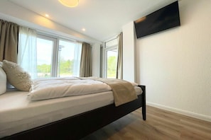 A modern bedroom with a bed, curtains, a flat-screen TV, and a view of the outdoors. - Holiday apartment with 1 bedroom (Cuxhaven)