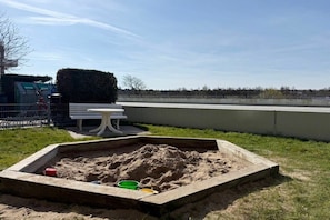A sandy play area with a bench and a picnic table, surrounded by a low wall and a clear sky. - Holiday apartment with 1 bedroom (Cuxhaven)