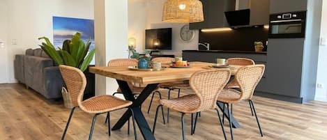 A modern dining area with a wooden table and wicker chairs, a television, and a kitchen in the background.