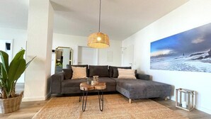 En moderne stue med en modulsofa, en vævet måtte, en hængende lampe og et stort landskabsmaleri på væggen.