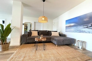 A modern living room with a sectional sofa, a woven rug, a hanging light fixture, and a large landscape painting on the wall. - Holiday apartment with 1 bedroom (Cuxhaven)