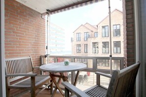 Uma varanda com uma mesa redonda e duas cadeiras de madeira, com vista para um edifício de tijolos e várias janelas.