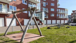 En legeplads med gyngestativ og rutchebane foran en høj bygning med altaner.