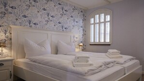 Un dormitorio con una cama grande, ropa de cama blanca, una mesita de noche con una lámpara y una ventana con persianas.