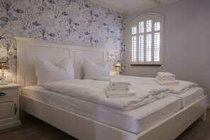 A bedroom with a large bed, white bedding, a nightstand with a lamp, and a window with blinds. - Holiday apartment in Büsum with swimming pool (Büsum)